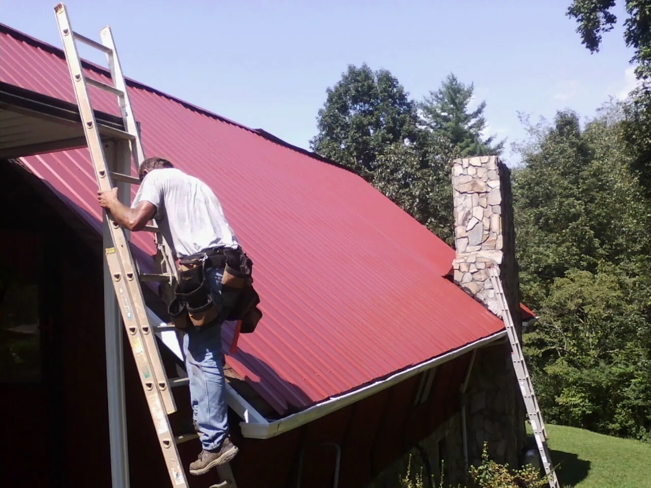 Roofer on a ladder beside a red metal roof and stone chimney — Soffit & Fascia Repair by Roofing Brothers in Mineral Point